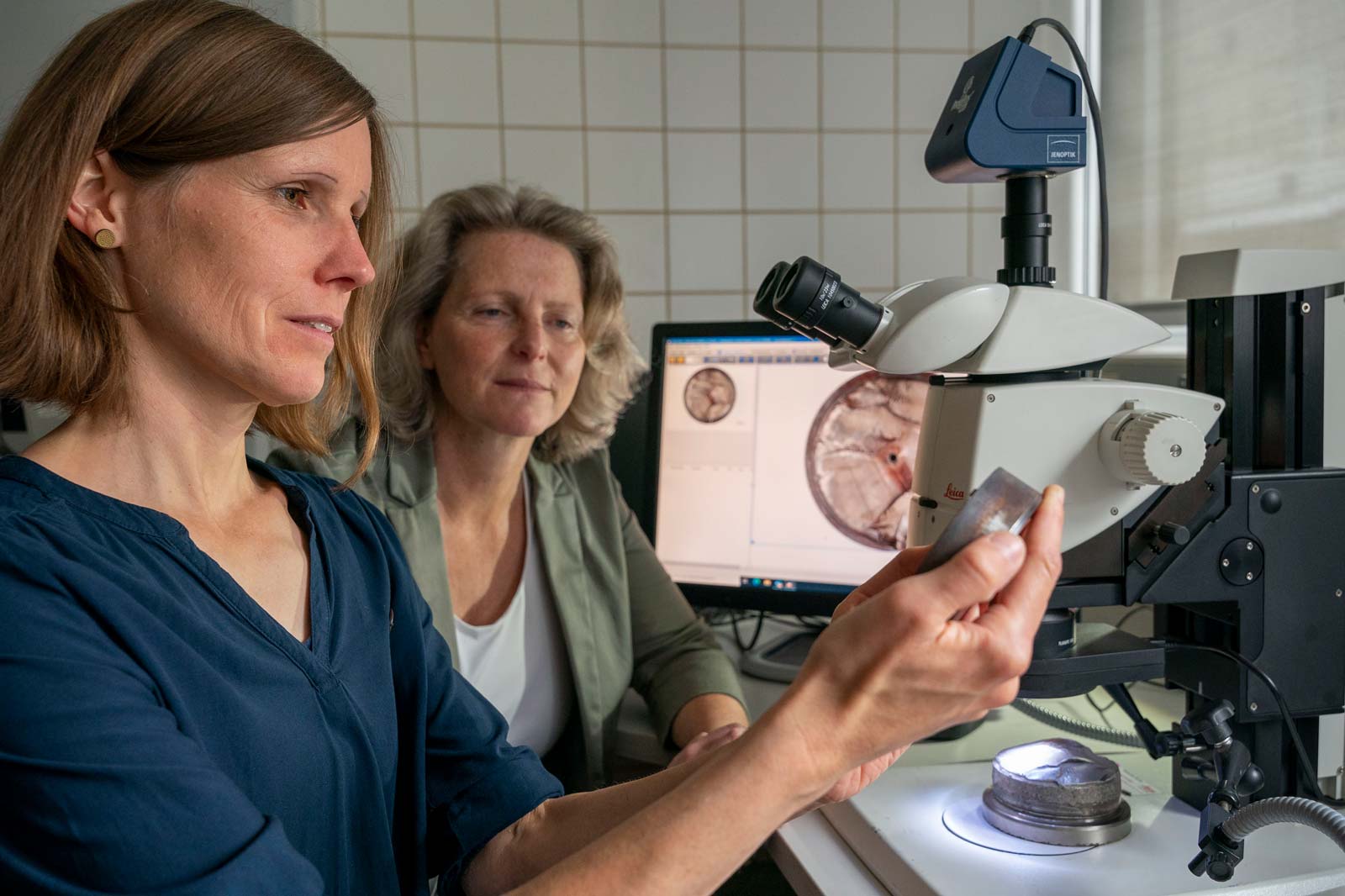 Zwei Frauen arbeiten an einem Mikroskop, während ein Computerbildschirm im Hintergrund sichtbar ist.