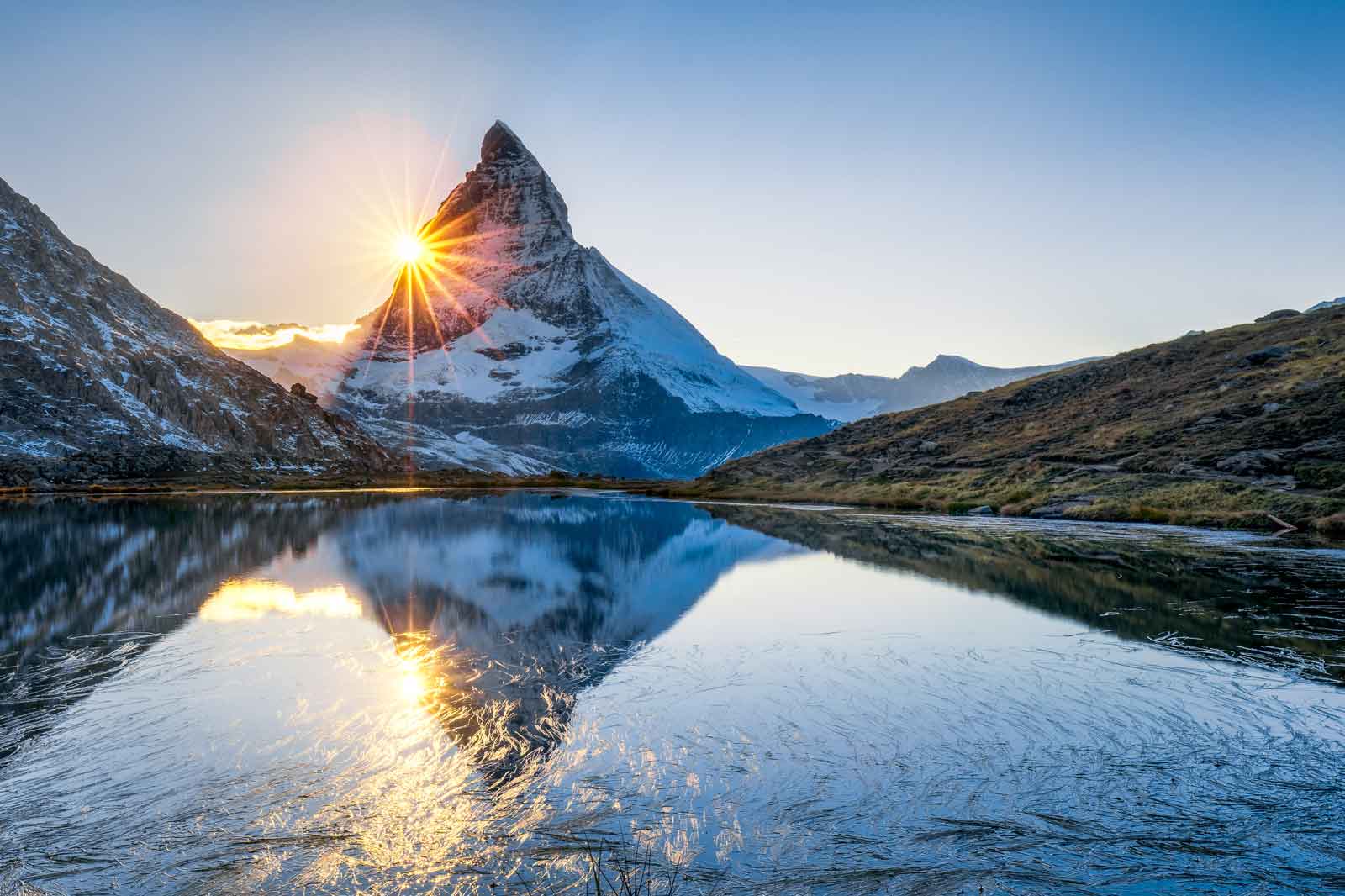 The Matterhorn mountain rises majestically in the background. Sun rays burst over the peak and reflect in the clear lake.