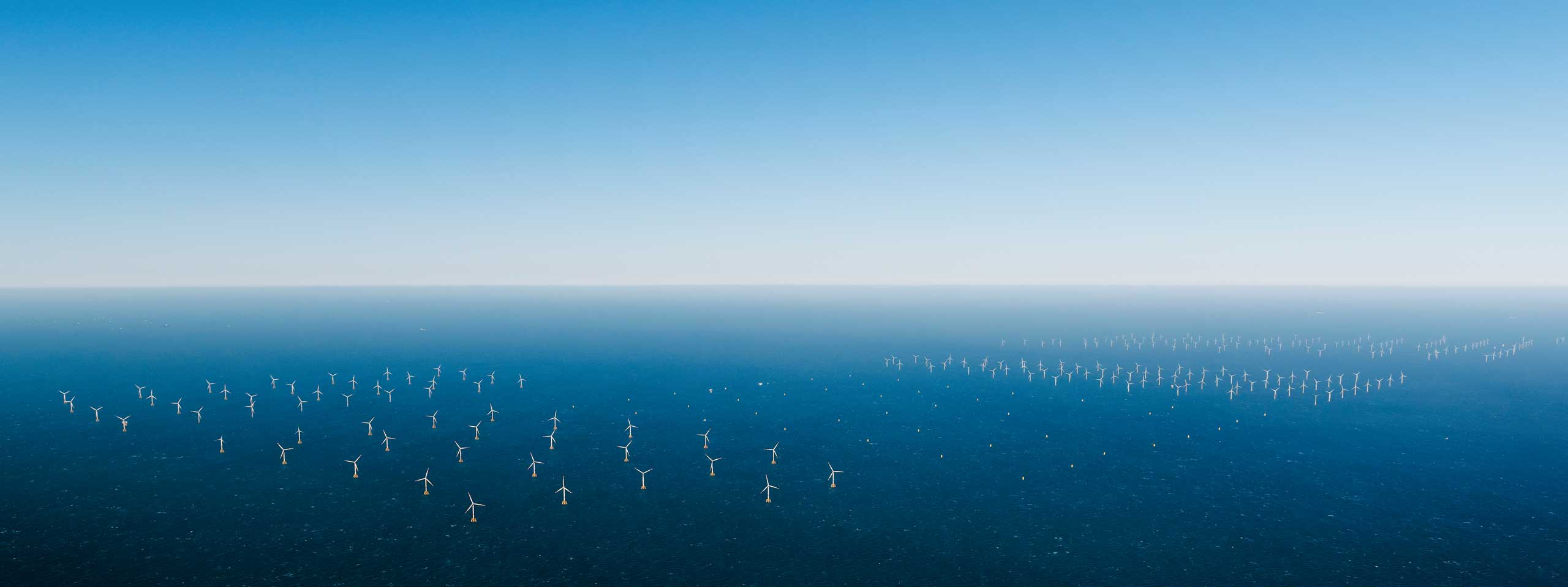 Eine weite Aussicht auf Offshore-Windkraftanlagen in einem ruhigen blauen Meer, unter einem klaren Himmel.