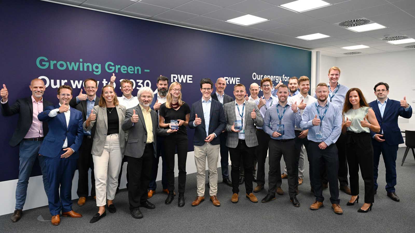 A group of people posing in an RWE office, giving thumbs up. A green slogan is visible in the background.