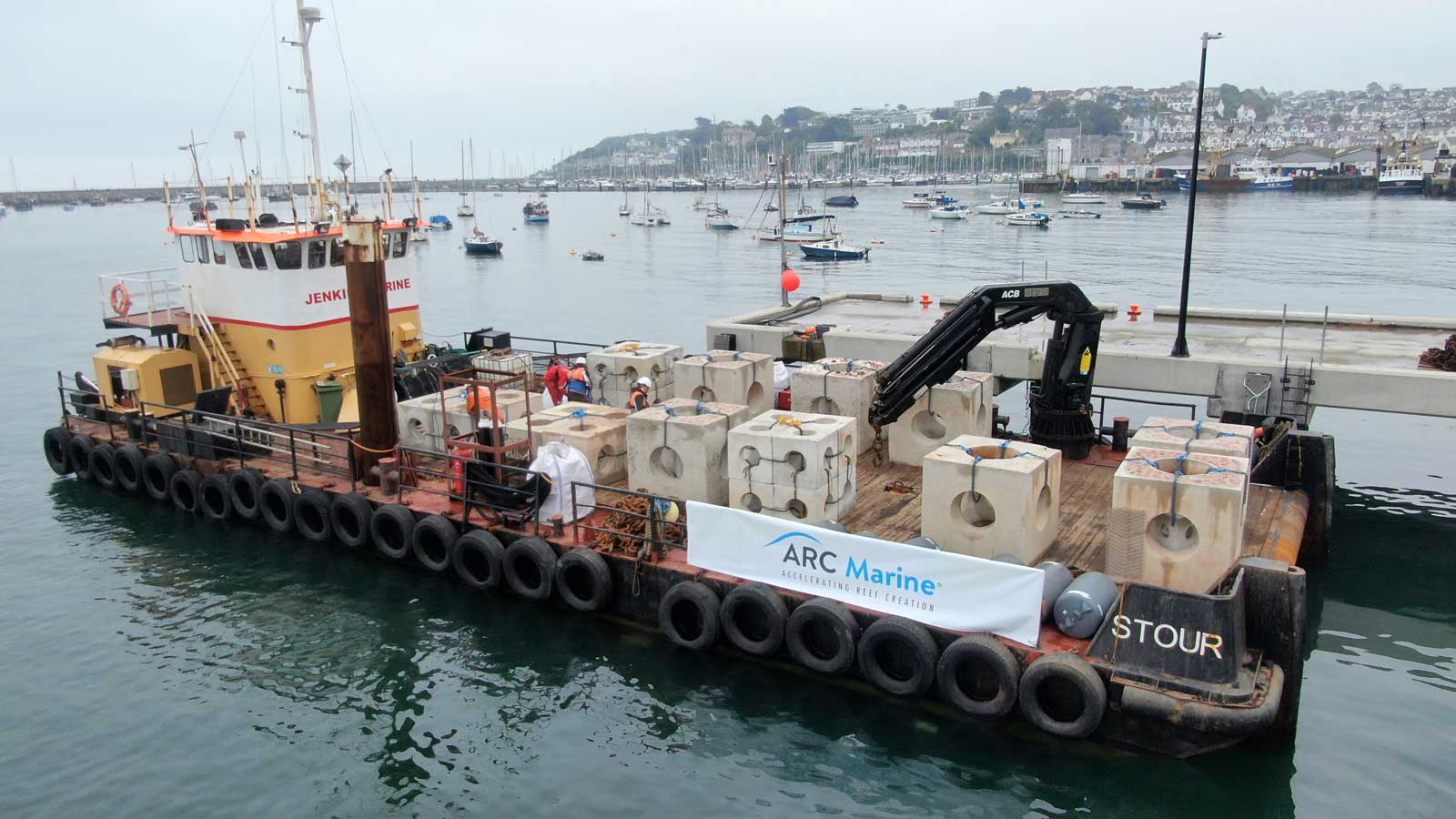 A barge named JENKINS MARINE loaded with concrete blocks is docked in a calm harbour with various boats in the background.