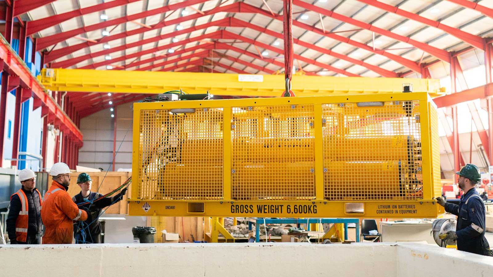 Ein gelbes Hebegerät wird in einer Fabrikhalle von mehreren Arbeitern bedient. Die Halle hat ein rotes Dach und helle Beleuchtung.