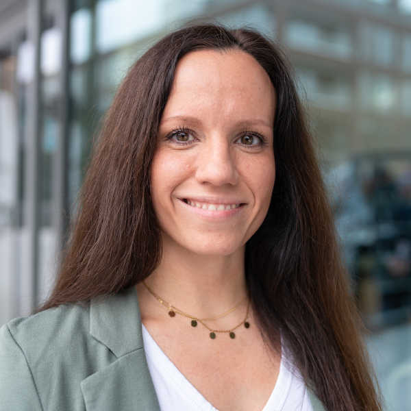 Eine Person mit langen, braunen Haaren trägt eine graue Jacke und eine weiße Bluse. Sie steht vor einem modernen Gebäude.