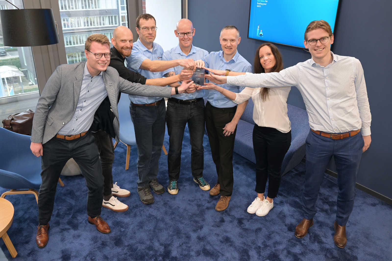 Eine Gruppe von Personen steht in einem modernen Raum und hält gemeinsam einen Preis, während sie in die Kamera lächeln.