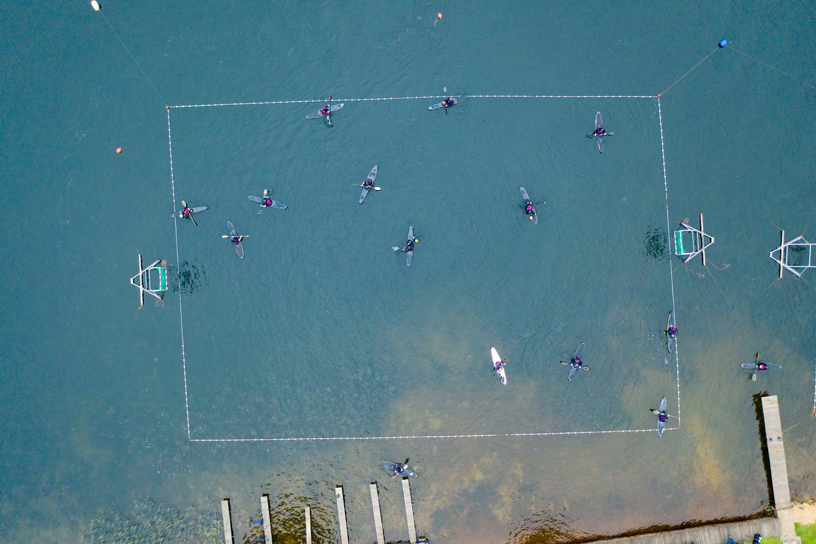 Eine Vogelperspektive auf Kajakfahrer in einem rechteckigen Bereich auf dem Wasser, mit einem Holzsteg im Vordergrund.