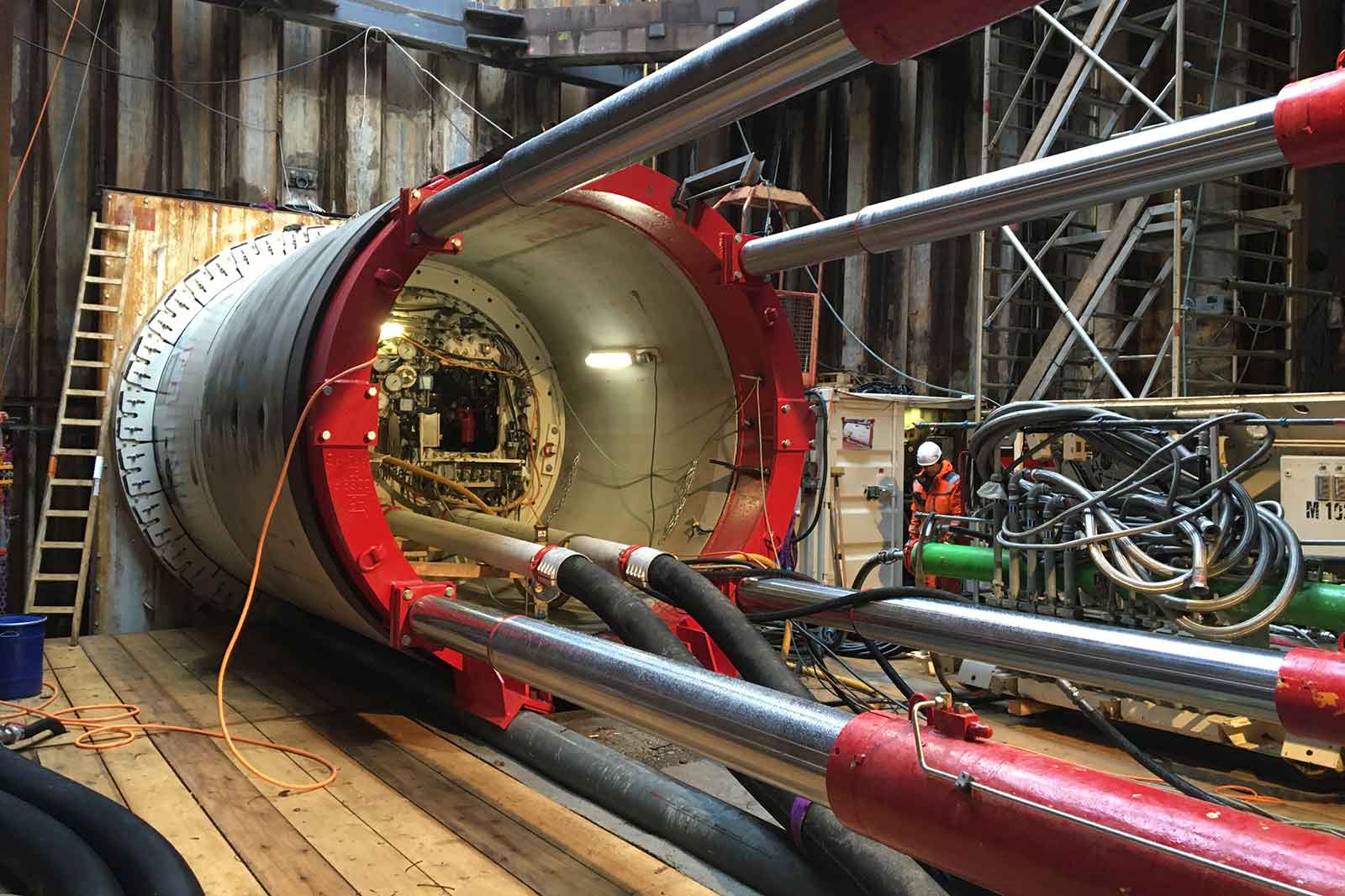 Großes zylindrisches Rohr mit rotem Rahmen, mehreren verchromten Hydraulikstangen, schwarzen Schläuchen, Kabeln, Holzboden, Leiter und einem Arbeiter in orangefarbener Warnkleidung und Helm.