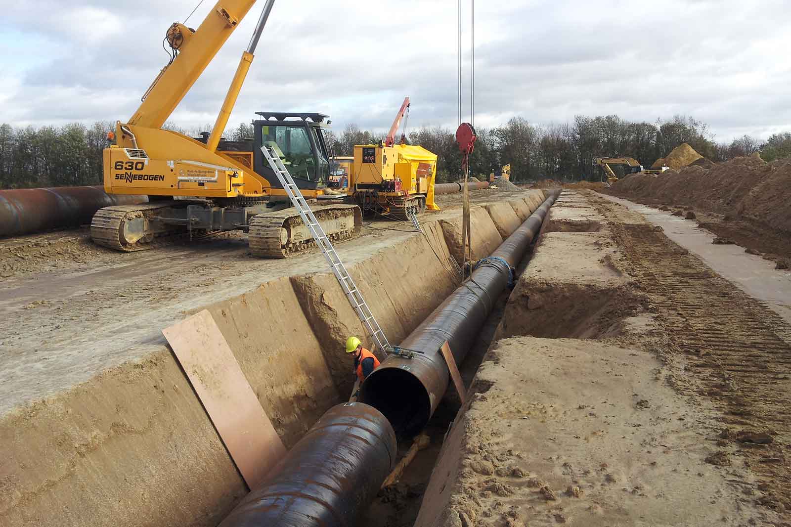Eine Baustellenszene mit einem tiefen Sandgraben, einer großen Stahlleitung, einem gelben Kran, Schweißgerät, Leiter und einem Arbeiter mit Schutzhelm.