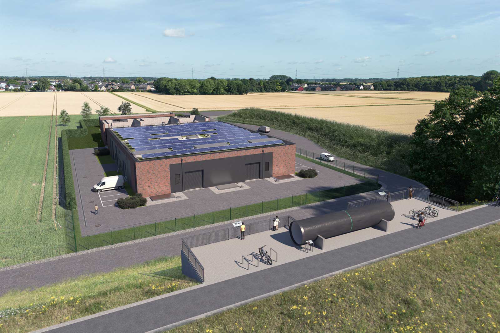 Ein rotes Backsteingebäude mit Solarpaneelen auf dem Dach steht neben Ackerland; ein eingezäunter Vorplatz, eine große schwarze Röhre, Fahrradständer und Personen sind zu sehen.