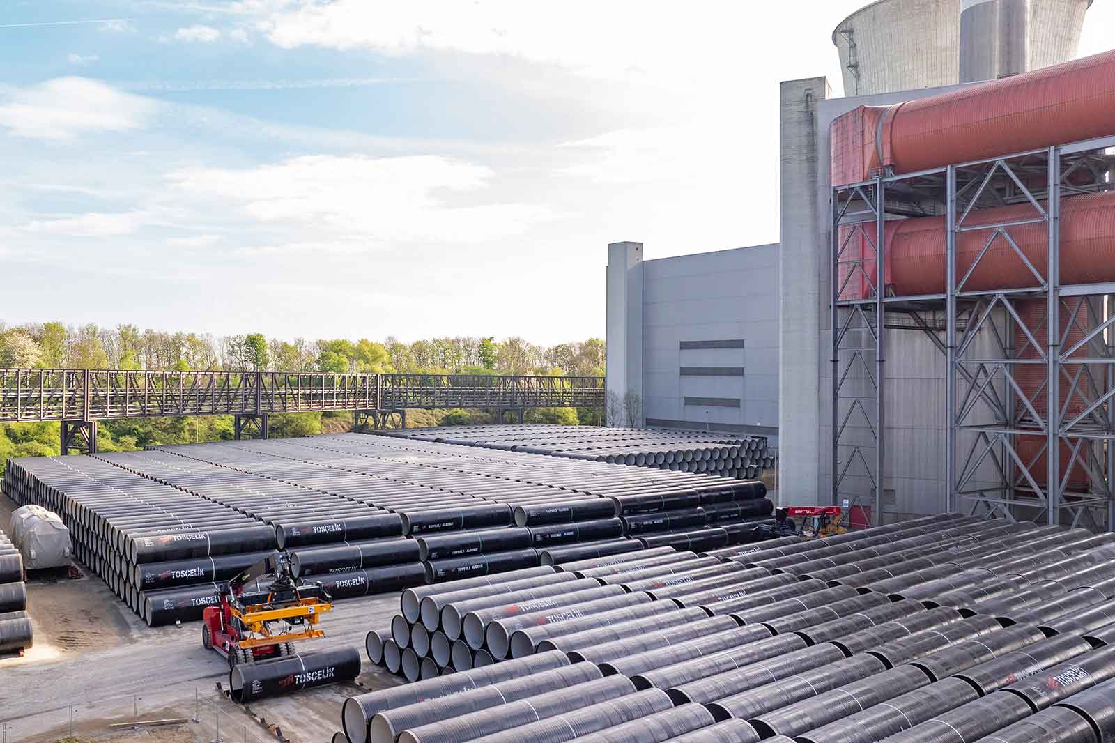 Großes Freilager mit ordentlich gestapelten schwarzen Stahlrohren neben einem Industriegebäude, mit sichtbaren roten Rohrleitungen und Hebegeräten.
