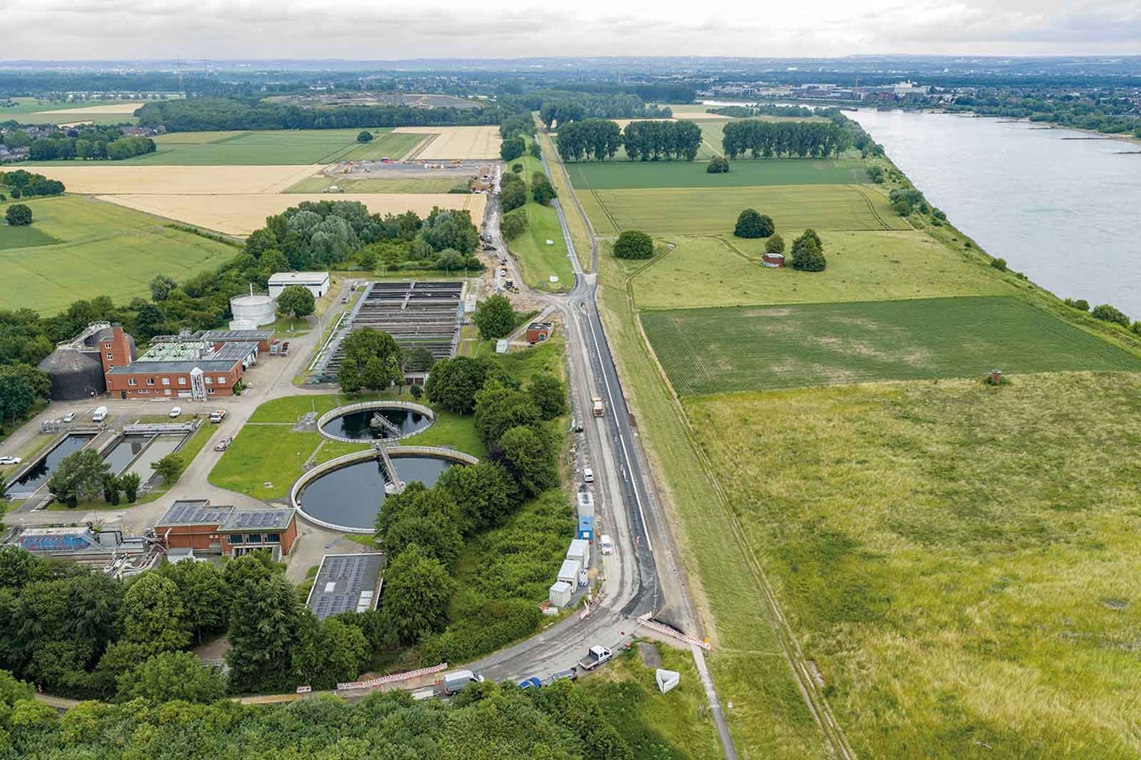 Luftaufnahme einer Kläranlage mit grauen Beton-Absetzbecken links, einer schnurgeraden Landstraße, grünen Feldern und einem breiten Fluss unter bewölktem Himmel.