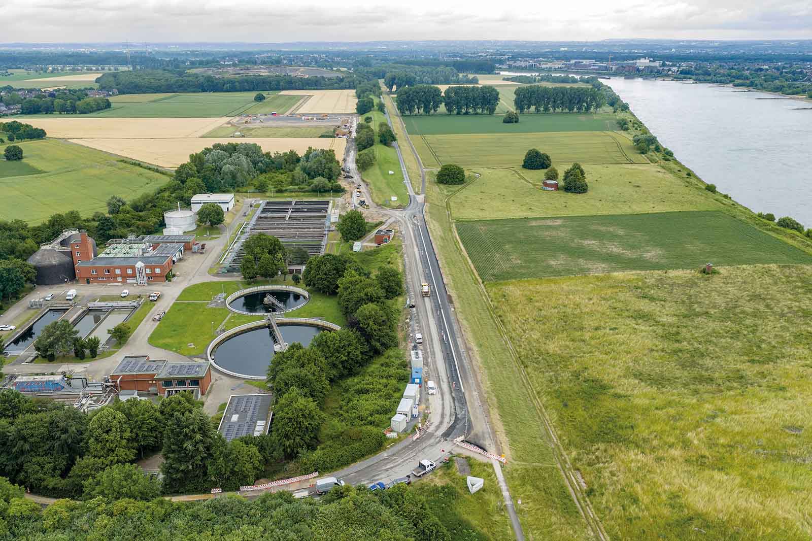Luftaufnahme einer Kläranlage mit grauen Beton-Absetzbecken links, einer schnurgeraden Landstraße, grünen Feldern und einem breiten Fluss unter bewölktem Himmel.