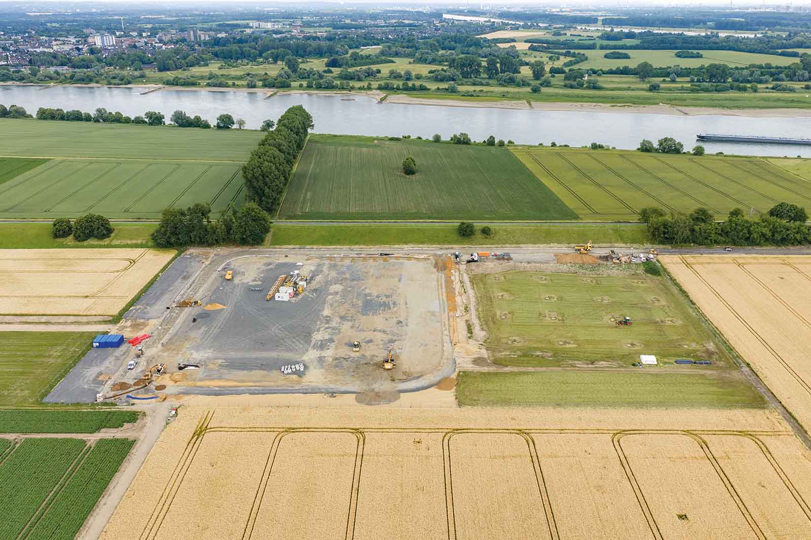 Im Vordergrund liegen goldene Getreidefelder; dahinter erstreckt sich eine großflächige Baustelle mit Schotterflächen und Baumaschinen, im Hintergrund ein Fluss und grüne Felder.