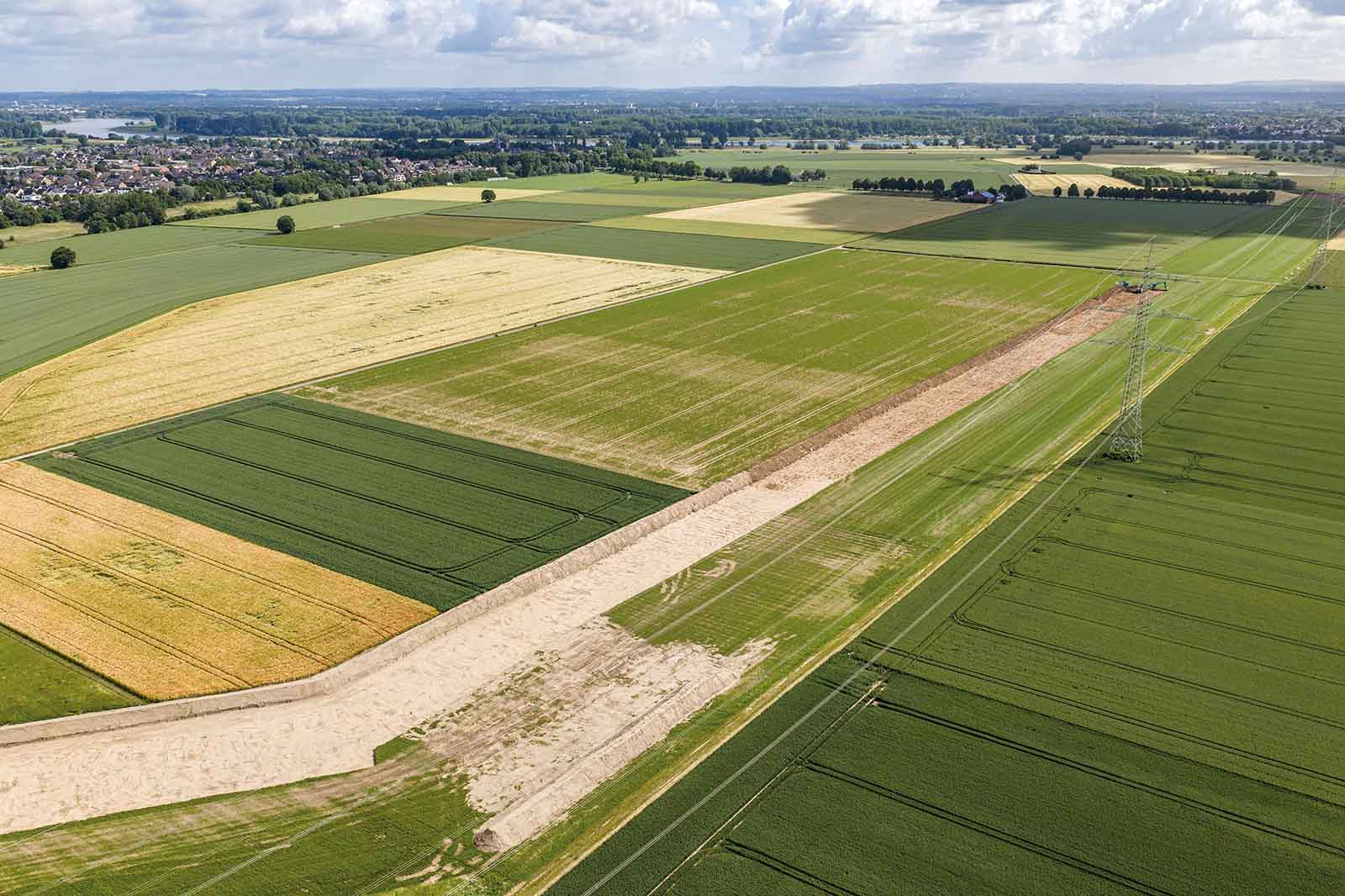 Blick aus der Luft auf rechteckige Ackerflächen in verschiedenen Grüntönen und goldgelbem Getreide, ein heller Sandweg und ein Strommast rechts, dahinter Dorf und bewölkter Himmel.
