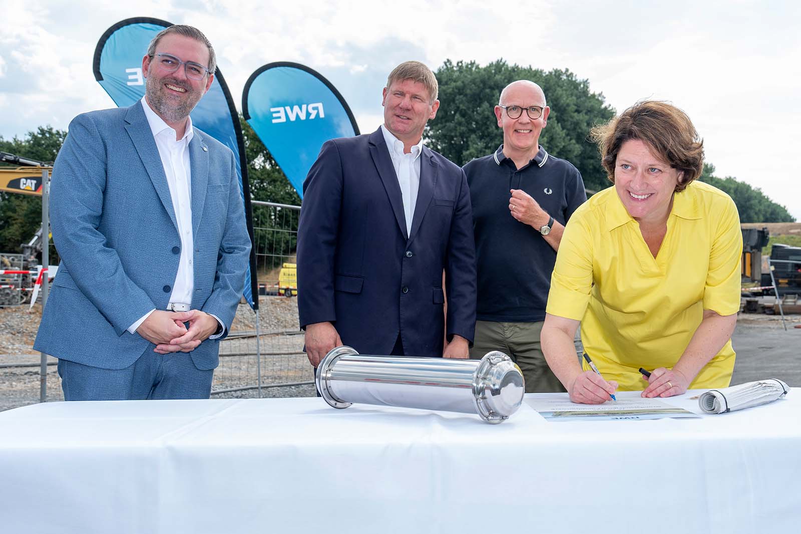 Vier Personen an einem weißen Tisch bei einer Baustelle; eine Frau in Gelb unterschreibt neben einer silbernen Kapsel, im Hintergrund blaue Wimpel mit „RWE“.