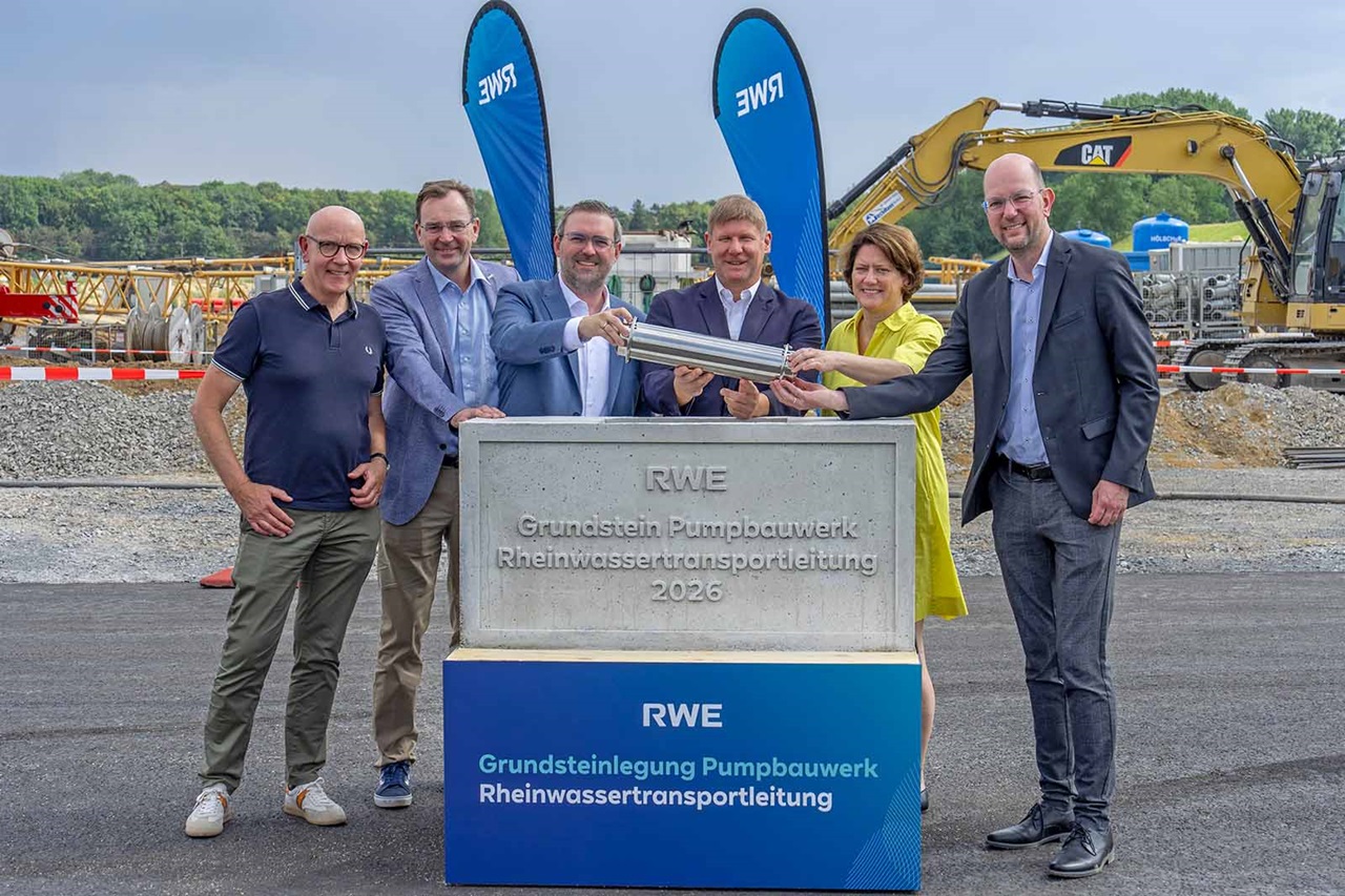 Sieben lächelnde Personen halten eine silberne Metallrolle vor einer Steinplatte mit den Aufschriften „RWE Grundstein Pumpwerk Rheinwassertransportleitung 2026“ und „RWE Grundsteinlegung Pumpwerk Rheinwassertransportleitung“.