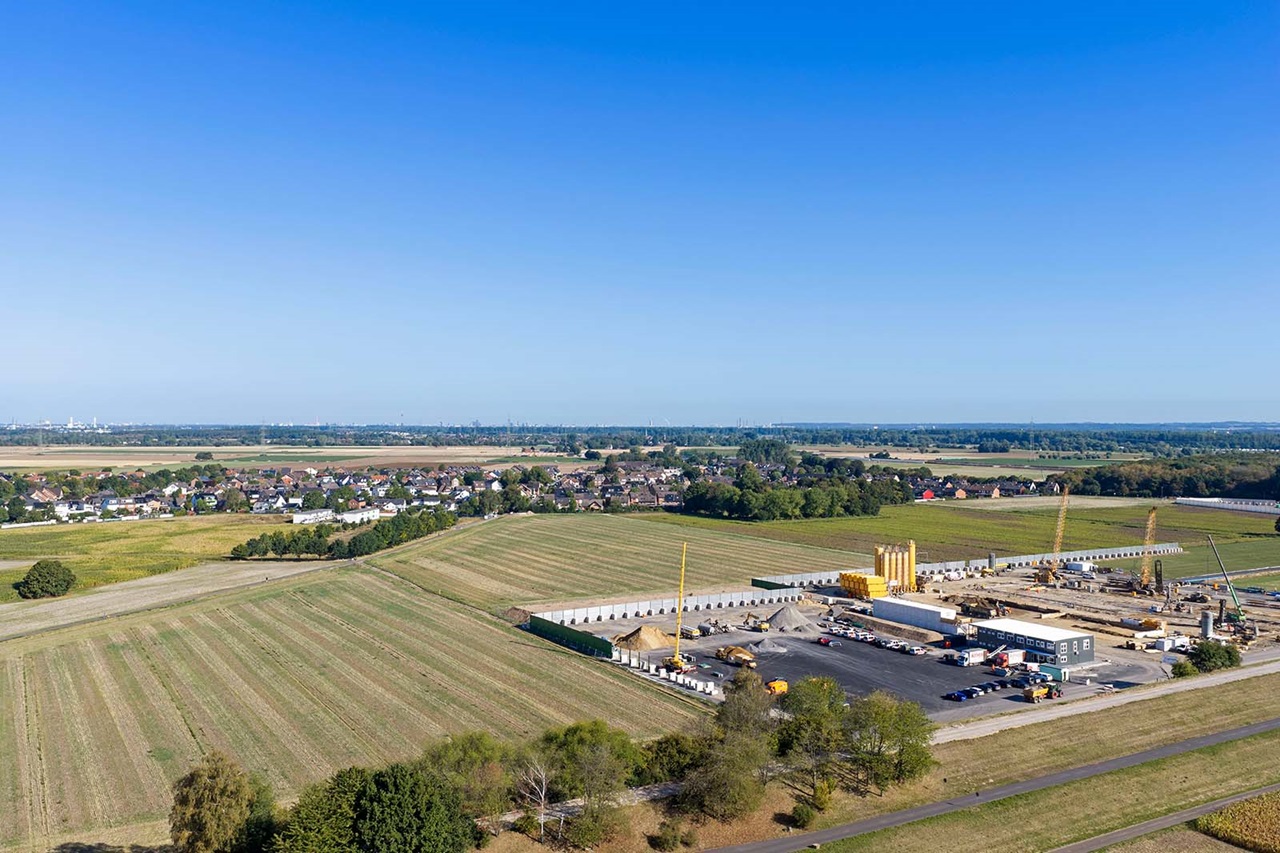 Im Vordergrund eine Baustelle mit gelben Silos, Kränen und abgestellten Fahrzeugen auf grauem Beton, umgeben von grünen Feldern und einem Dorf unter klarem blauen Himmel.