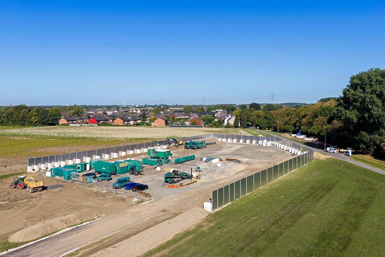 Eine temporäre Baustelle mit grünen Maschinen und Containern auf hellbraunem Erdgrund, eingerahmt von weißen Stellwänden, Rasenböschung und roten Backsteinhäusern unter klarem blauem Himmel.