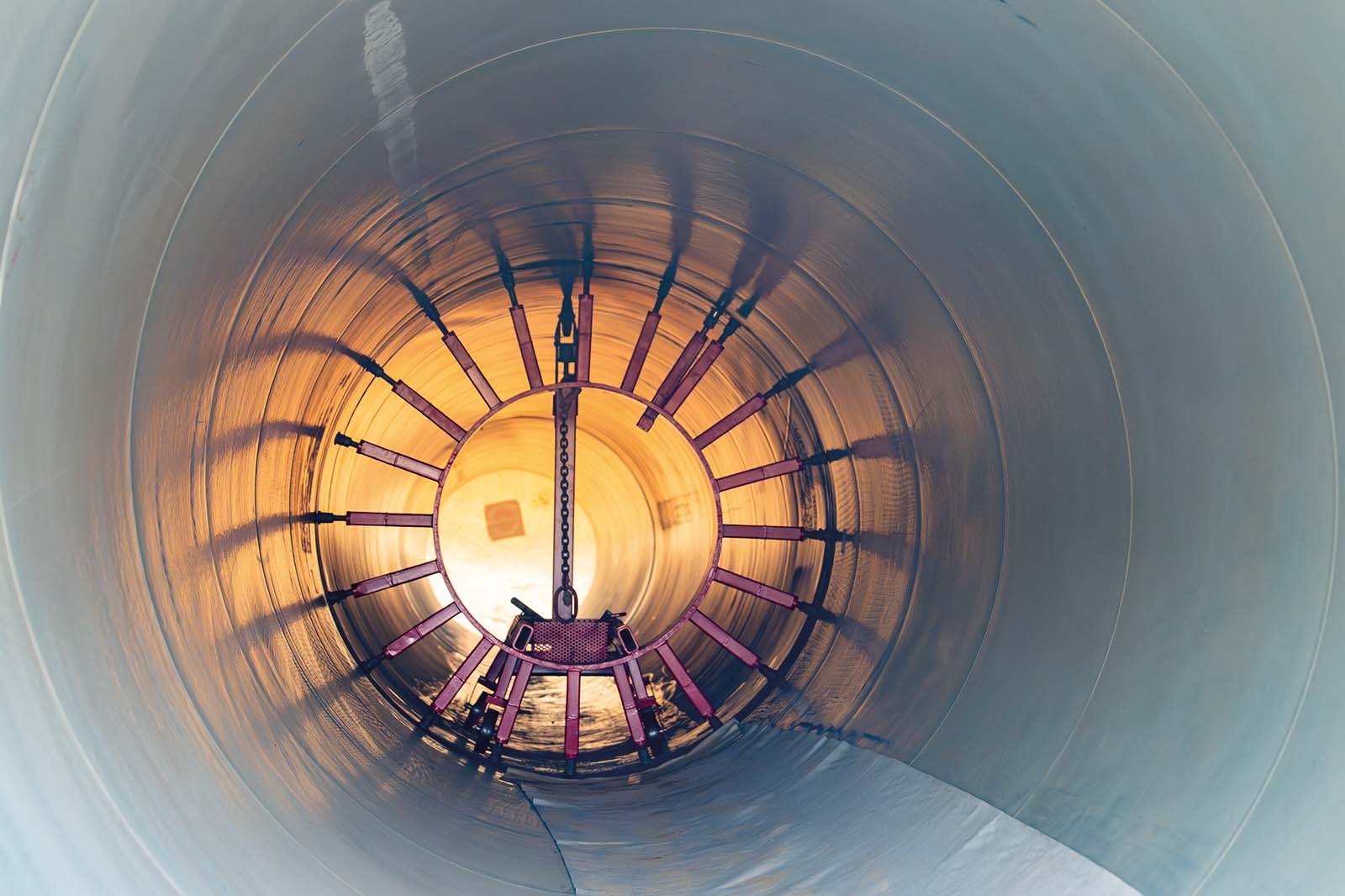 Blick in das Innere eines großen zylindrischen Metallrohres mit radialen Streben und einer kettengeführten Mechanik vor einer warm goldenen Öffnung.