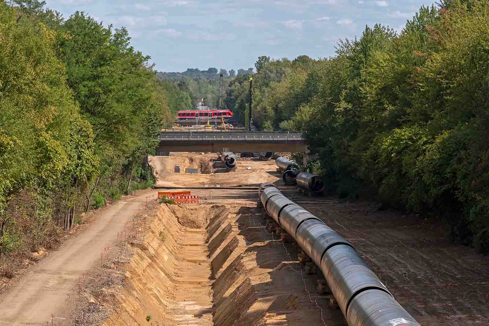 Große schwarze Stahlrohre liegen in einer sandigen Baugrube, flankiert von grünen Bäumen; im Hintergrund fährt ein roter Zug über eine Betonbrücke, die Szene wirkt industriell.