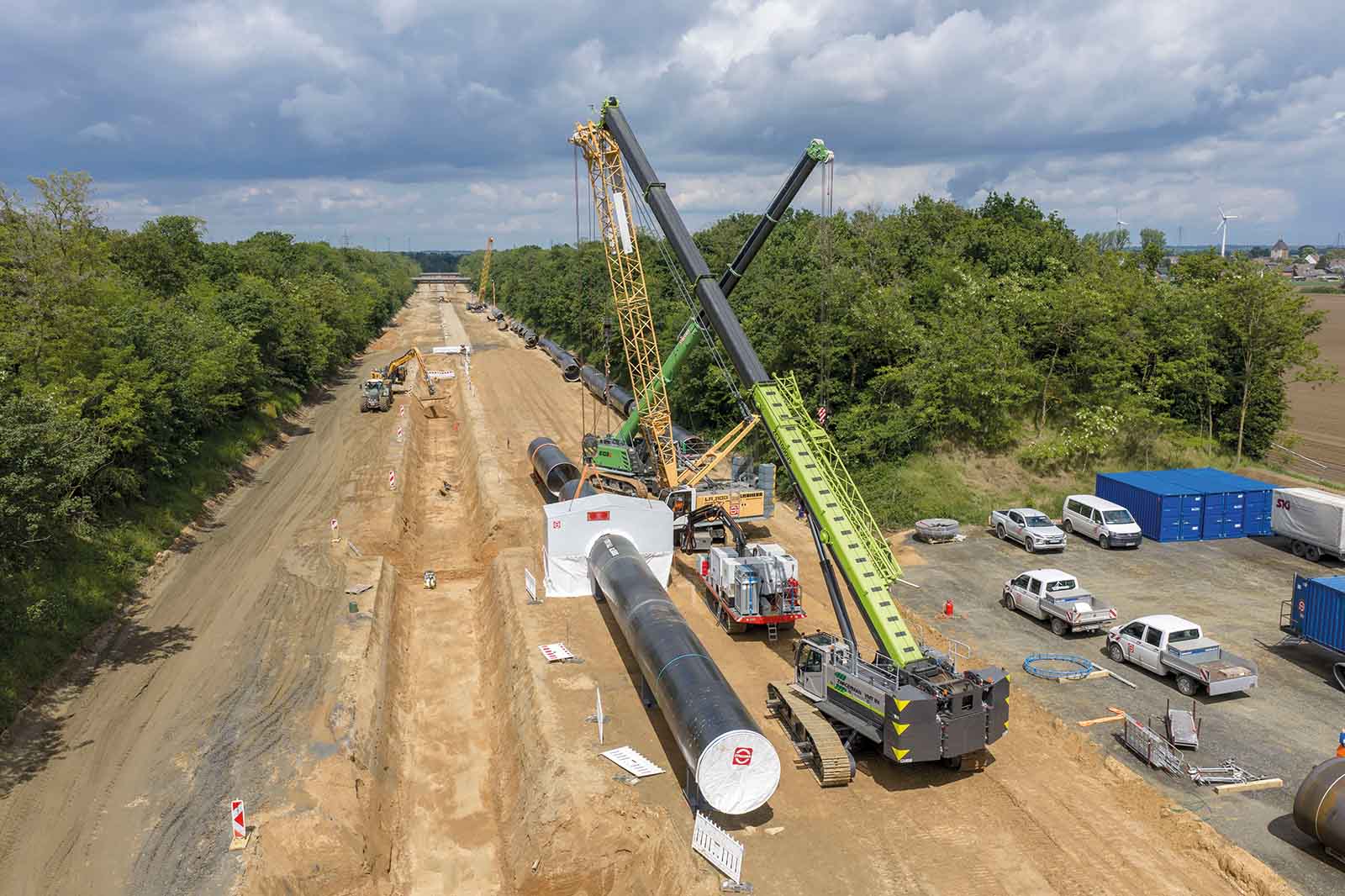 Zwei große Kräne heben ein großes schwarzes Rohrsegment über einem breiten, sandigen Graben auf einer Pipeline-Baustelle, umgeben von Bäumen und mehreren weißen Transportern.