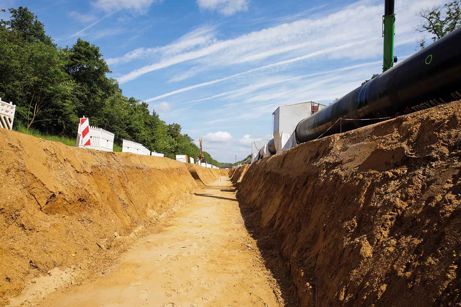 Eine tiefe sandfarbene Baugrube mit losem Erdweg; rechts verläuft eine schwarze Rohrleitung, links stehen weiße Baucontainer und Bäume unter blauem Himmel.