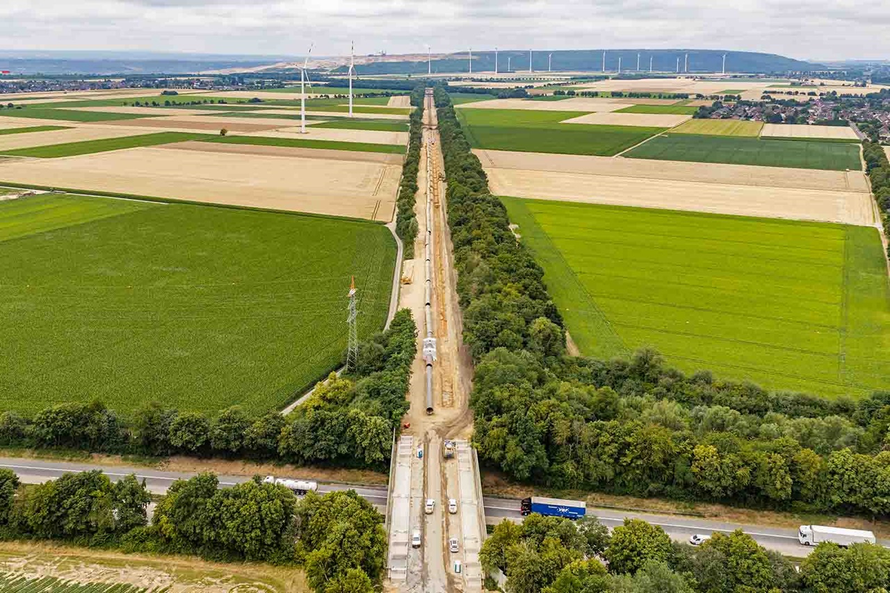 Eine lange, gerade Baustelle mit mehreren Fahrzeugen und freigelegtem Erdstreifen verläuft mittig durch grüne und gelbe Ackerflächen, flankiert von Baumreihen und Windkraftanlagen am Horizont.