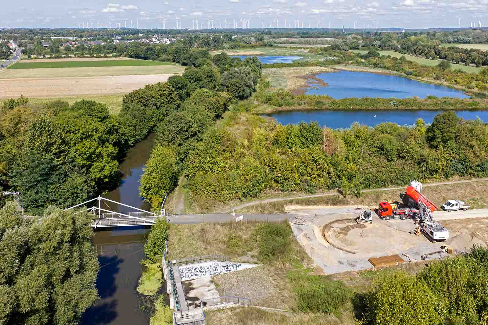 Ein Bauplatz mit rotem Betonmischer und weißem Transporter neben einem schmalen Weg und einer weißen Fußgängerbrücke, dahinter blaue Teiche und grüne Felder unter hellem Himmel.