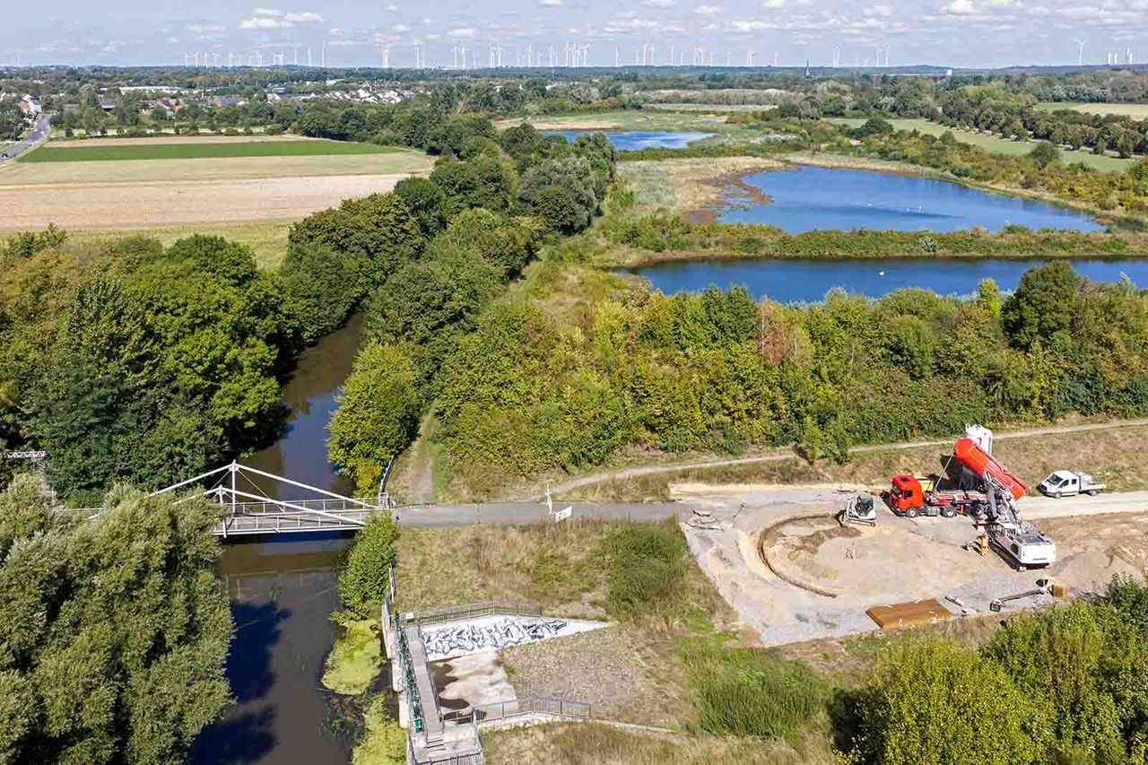 Ein Bauplatz mit rotem Betonmischer und weißem Transporter neben einem schmalen Weg und einer weißen Fußgängerbrücke, dahinter blaue Teiche und grüne Felder unter hellem Himmel.