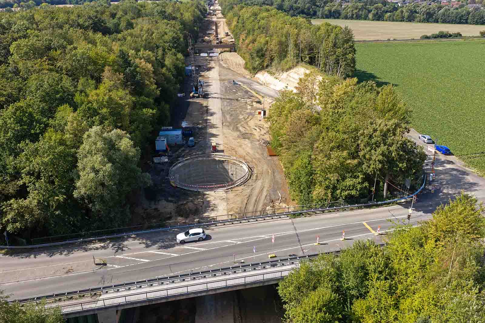 Eine weiße Limousine fährt über eine graue Betonbrücke im Vordergrund, dahinter eine braune Erdbaustelle mit rundem Betonfundament und grünen Feldern.
