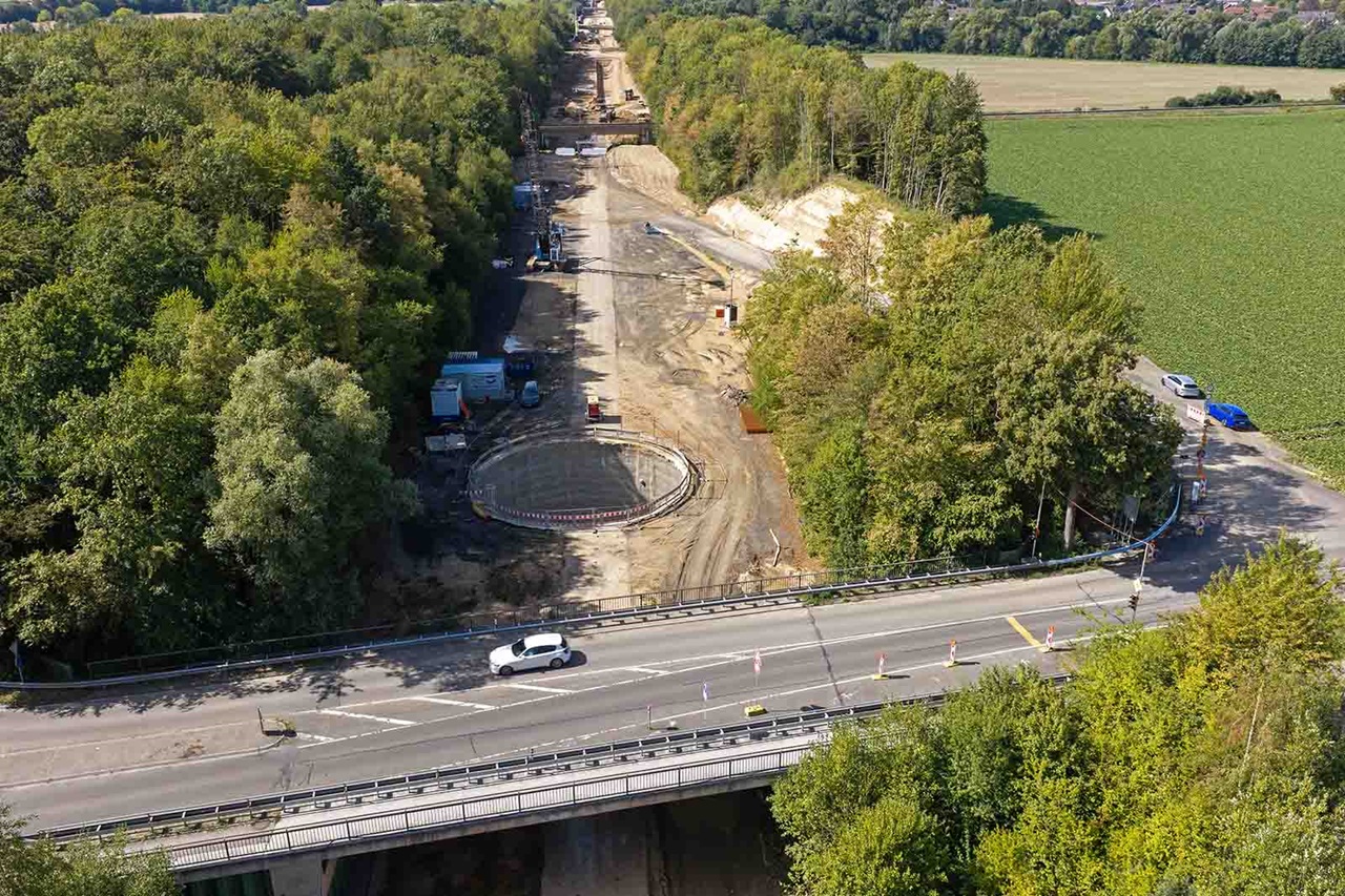 Eine weiße Limousine fährt über eine graue Betonbrücke im Vordergrund, dahinter eine braune Erdbaustelle mit rundem Betonfundament und grünen Feldern.