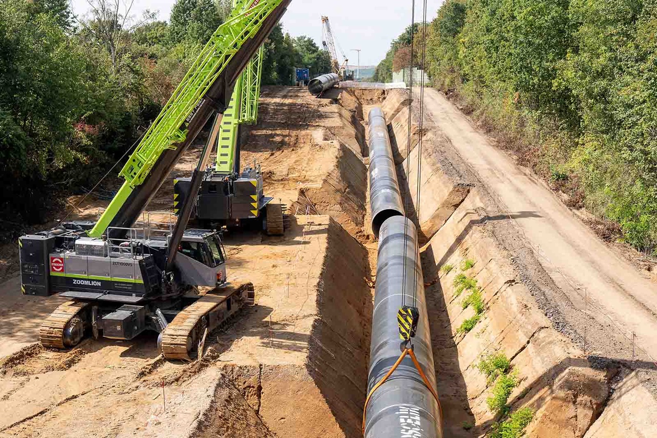 Ein grüner Kran mit Raupenfahrwerk hebt ein großes schwarzes Stahlrohr in einer tiefen sandigen Baugrube neben einer unbefestigten Straße und dichtem Baumbestand.