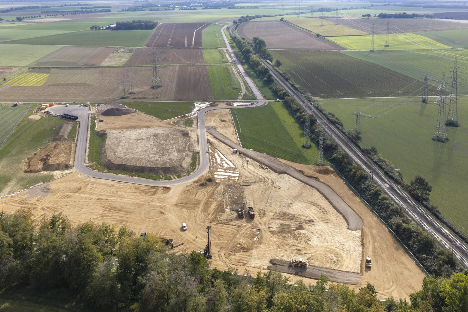 Luftaufnahme einer großflächigen Baustelle mit hellbraunem aufgefülltem Erdreich, mehreren Baufahrzeugen und neu angelegten Straßen, umgeben von grünen Feldern und Strommasten.