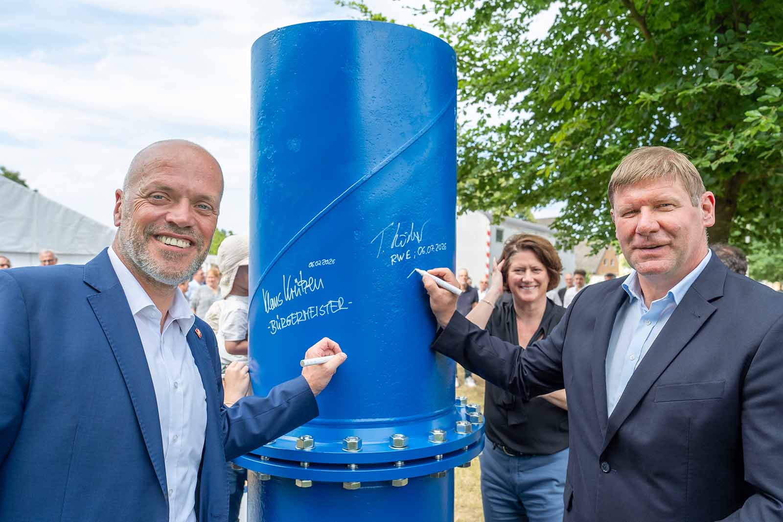 Zwei Männer in dunkelblauen Anzügen signieren mit weißen Markern eine hohe blaue Metallröhre mit sichtbaren Schrauben, hinter ihnen lächelt eine Frau im Freien.