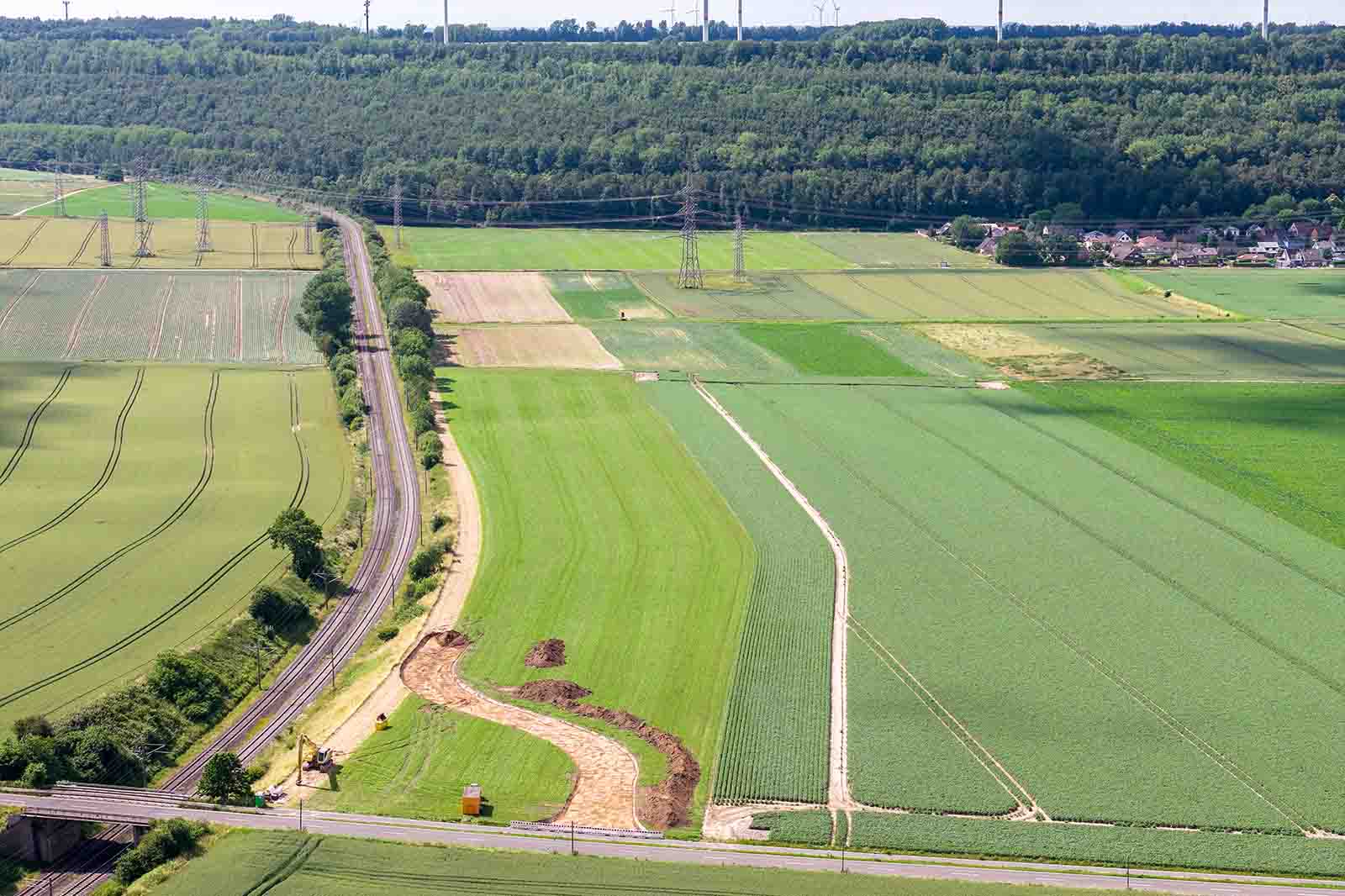 Eine Luftaufnahme zeigt weitläufige, verschieden grüne Ackerflächen mit einer geschwungenen Baumreihe links, verstreuten Hochspannungsmasten und einem bewaldeten Hügel im Hintergrund.