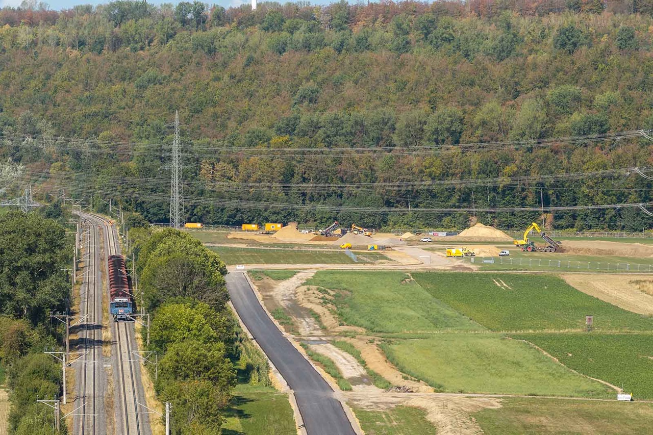 Ein roter Güterzug fährt links auf Bahngleisen entlang, rechts eine Baustelle mit gelben Baggern und Sandhügeln sowie frisch asphaltierter Straße vor einem bewaldeten Hügel.