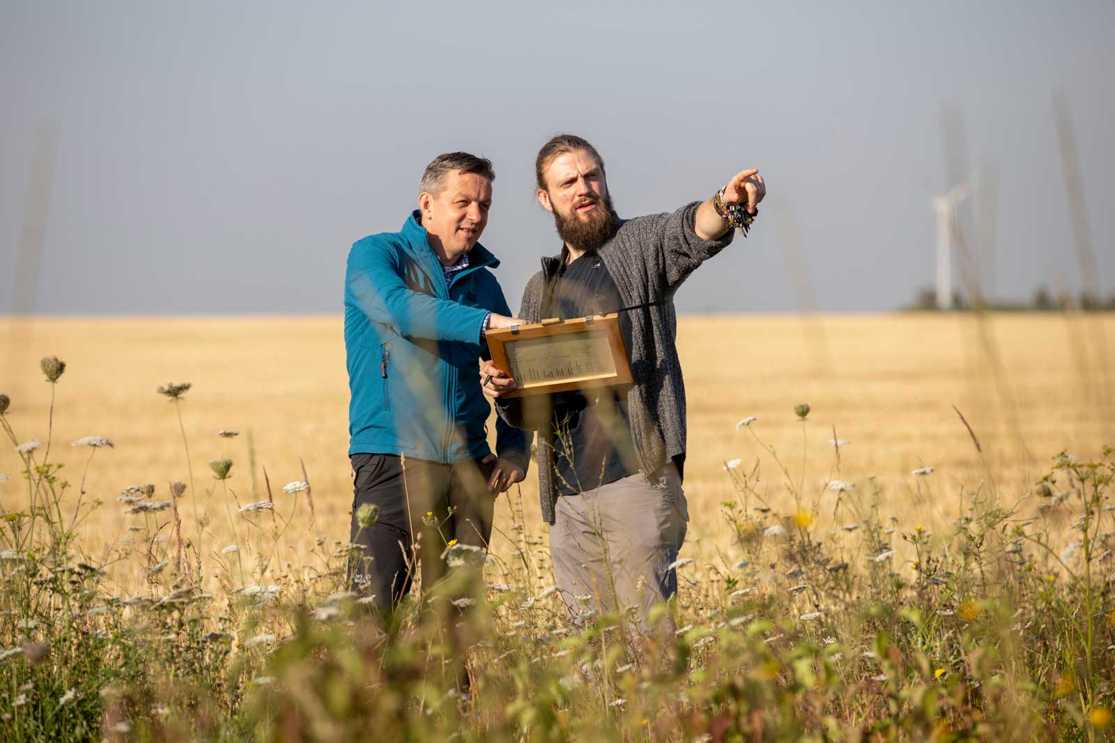 Zwei Männer in einem Feld, die einen Holzrahmen untersuchen, mit einem Windrad in der Ferne und Blumen um sie herum.