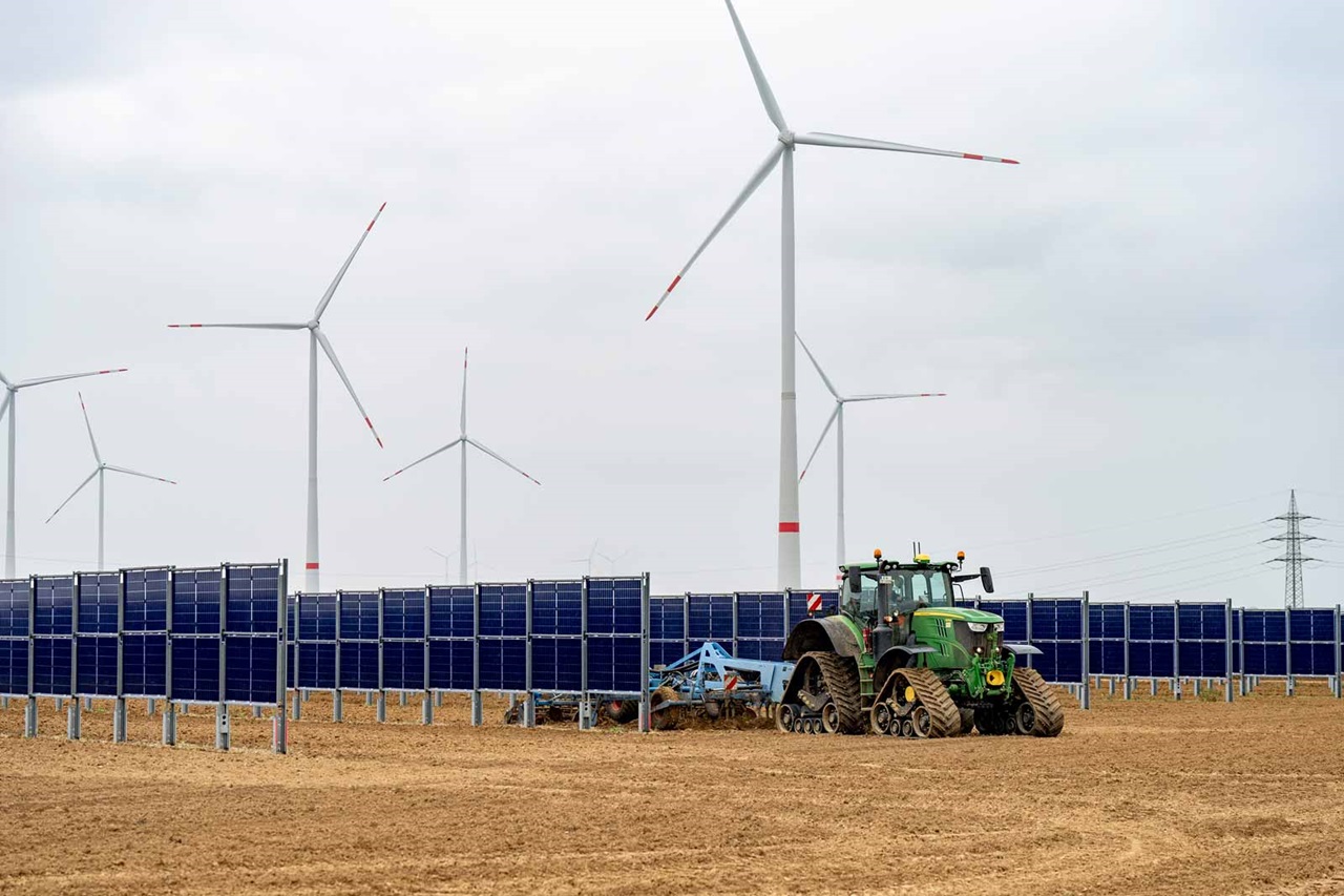 Ein Traktor pflügt zwischen Solarpanelen und Windrädern auf einem Feld unter bewölktem Himmel.