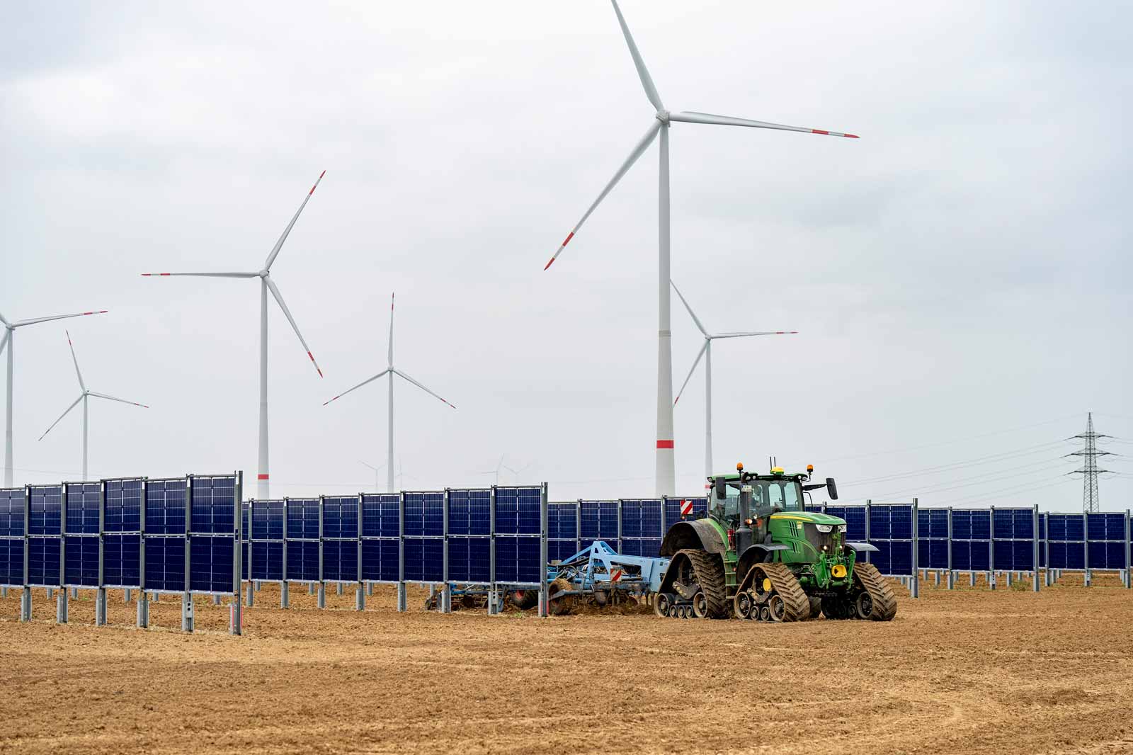 Ein Traktor pflügt zwischen Solarpanelen und Windrädern auf einem Feld unter bewölktem Himmel.