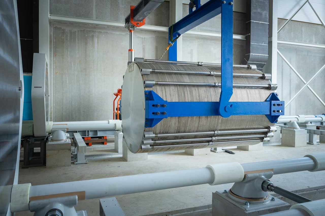 Ein großer Metallzylinder mit Drähten wird in einer industriellen Umgebung von einem blauen Kran angehoben.