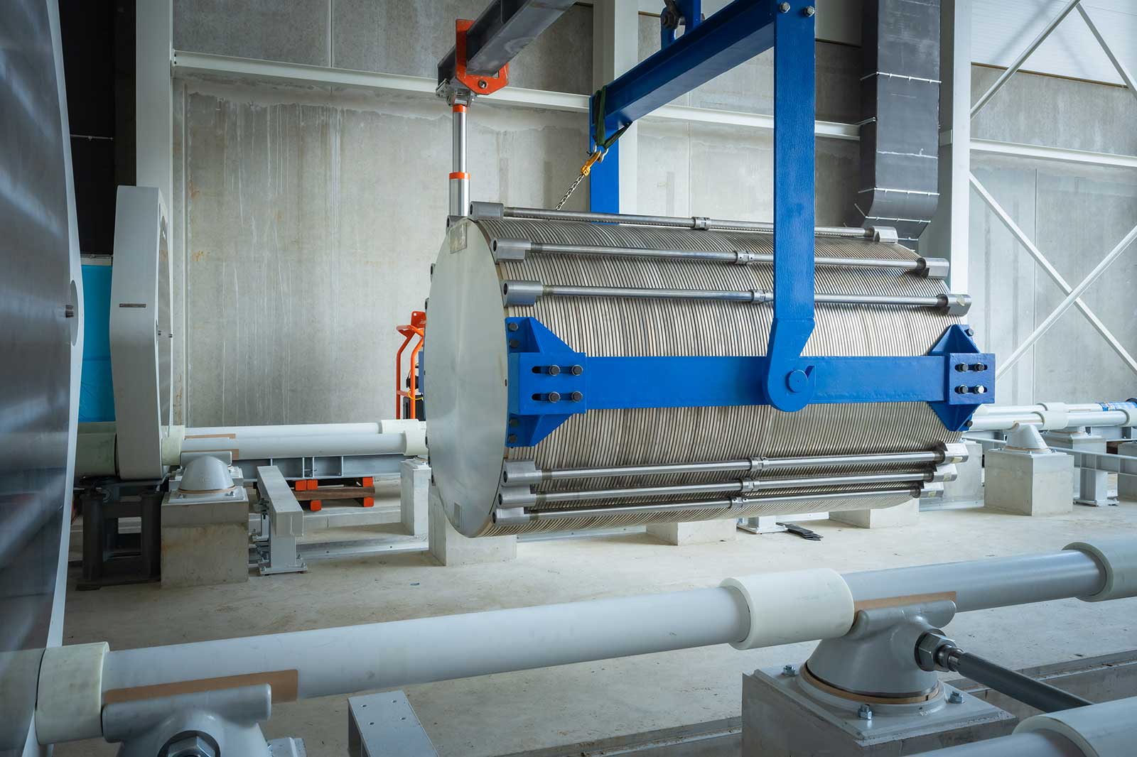 Ein großer Metallzylinder mit Drähten wird in einer industriellen Umgebung von einem blauen Kran angehoben.