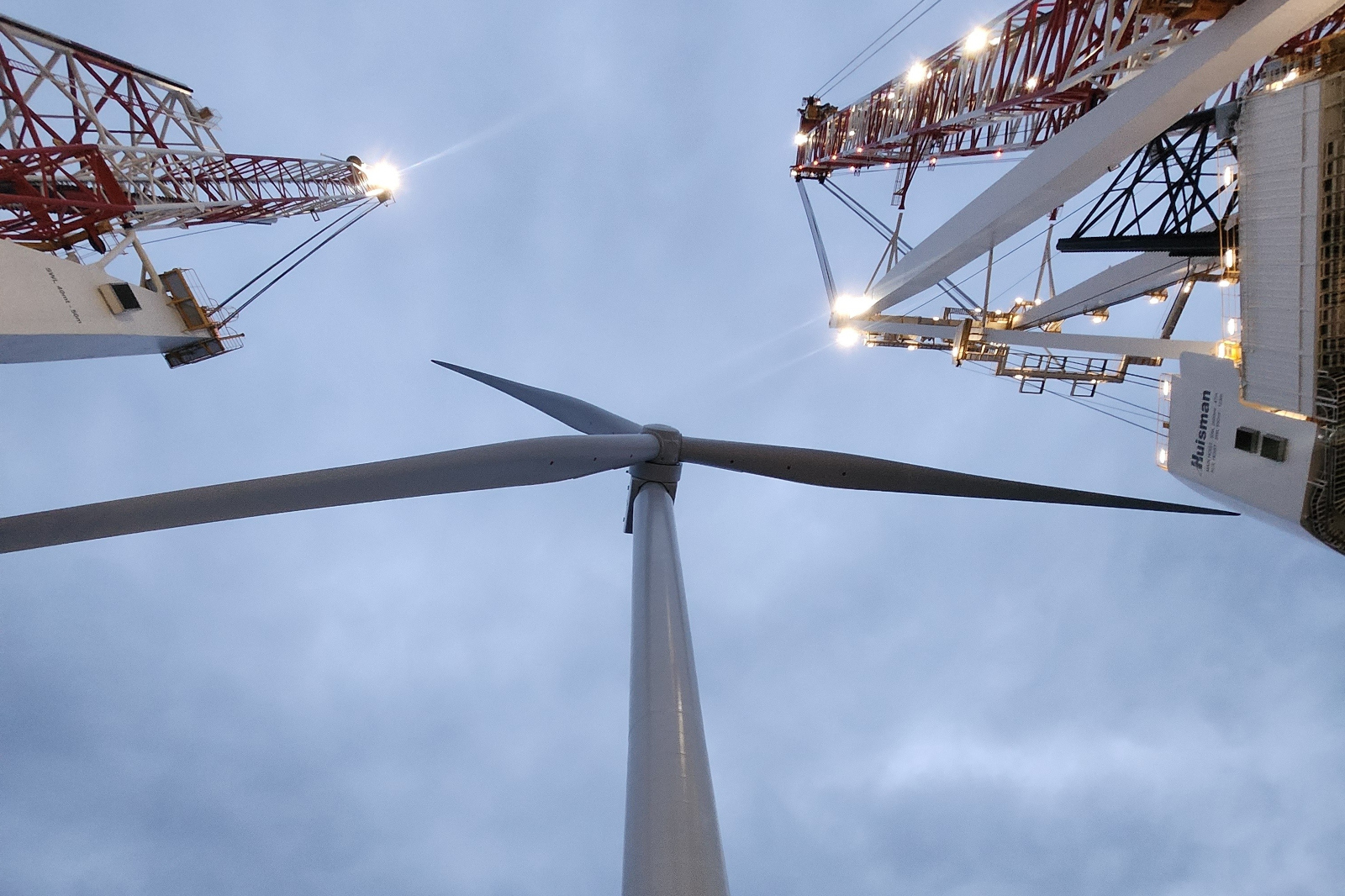A view from below of a wind turbine situated between two cranes, under a cloudy sky.
