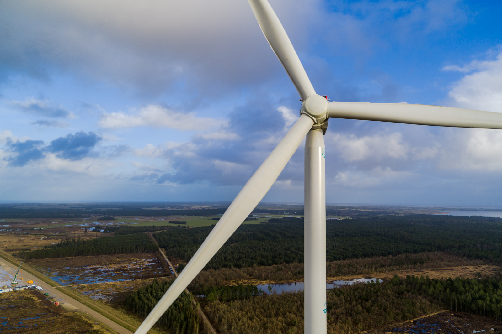 Close-up of a wind turbine against a blue sky with clouds. Fields and forests stretch out in the background.