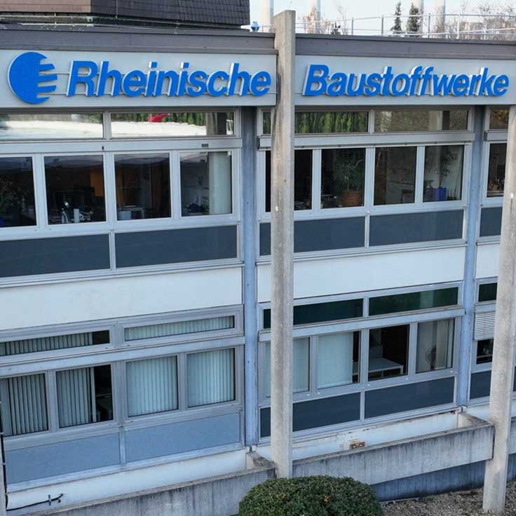 Die Außenansicht der Rheinischen Baustoffwerke zeigt ein modernes Gebäude mit großen Fenstern und einem blauen Schild.