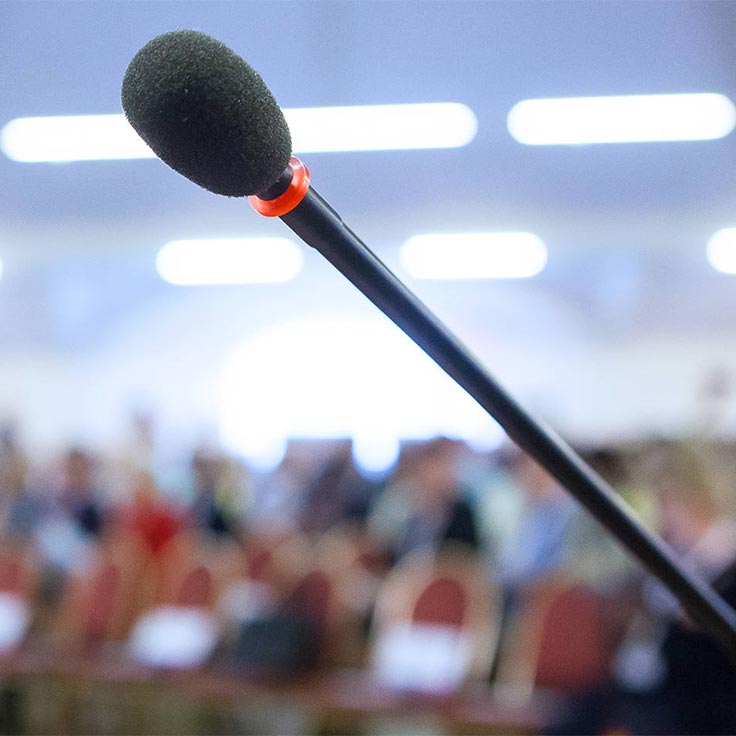 Ein Mikrofon auf einem Ständer ist im Fokus, mit einem verschwommenen Publikum im Hintergrund.