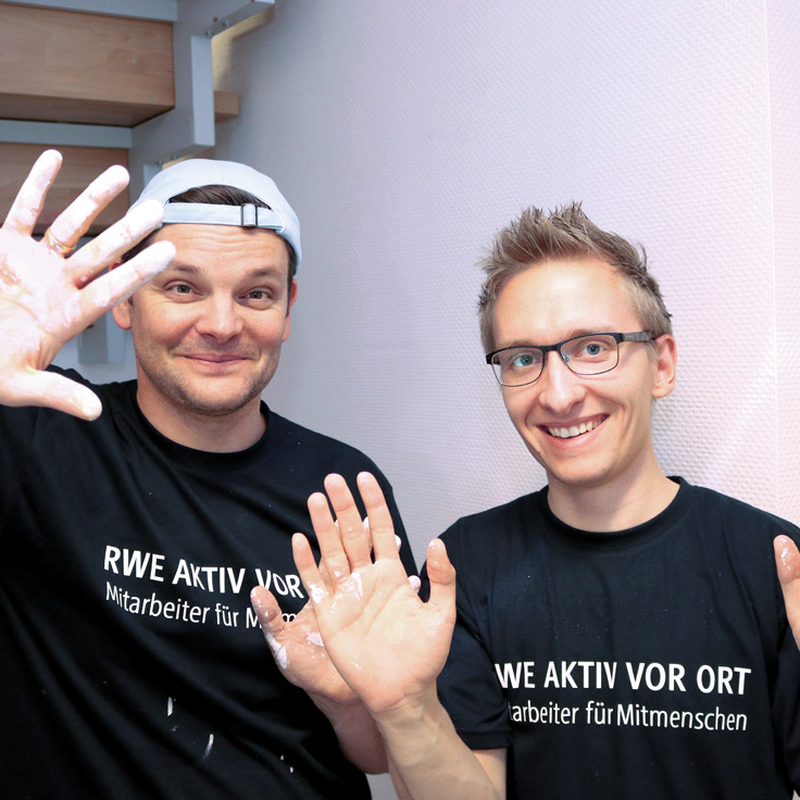 Zwei Männer zeigen ihre Hände, die mit weißem Kreide bedeckt sind. Sie tragen schwarze T-Shirts mit weißem Text.