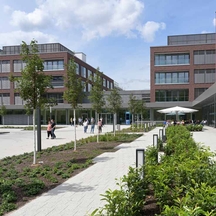 Ein moderner Campus mit Menschen, die in einem gestalteten Innenhof spazieren gehen, und Gebäuden im Hintergrund unter einem teilweisen bewölkten Himmel.