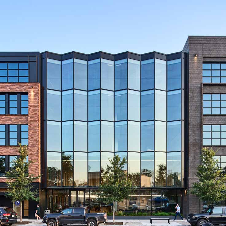 A modern building with a glass facade, surrounded by trees and a parking area in the foreground.