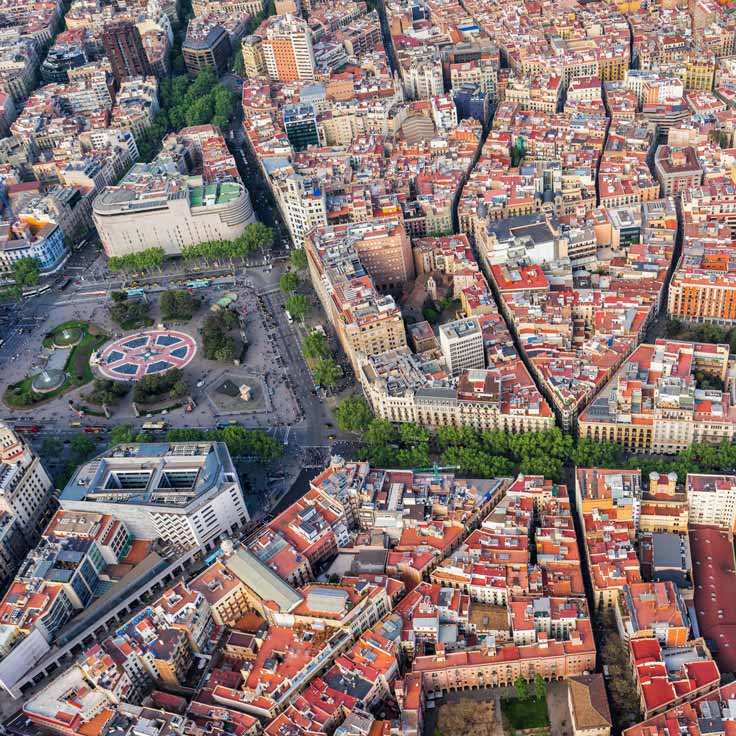 Luftaufnahme einer Stadt mit Häusern in verschiedenen Farben und einem zentralen Platz, umgeben von Bäumen.