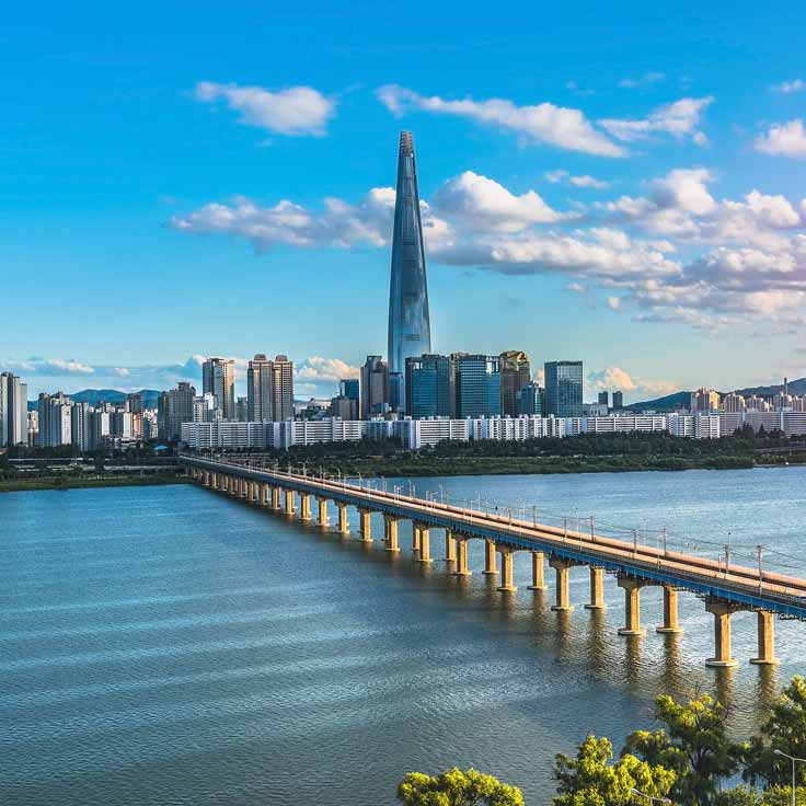 A skyline view featuring the Lotte World Tower and a bridge over water, under a clear blue sky with scattered clouds.