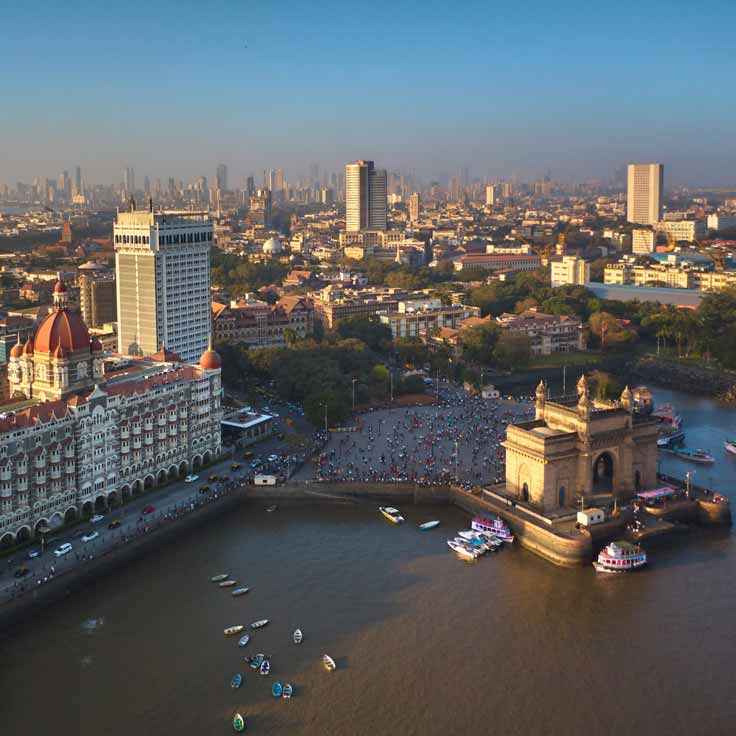 Luftaufnahme von Mumbai mit dem Gateway of India, umgeben von Stadtgebäuden und Menschenmengen am Wasser.